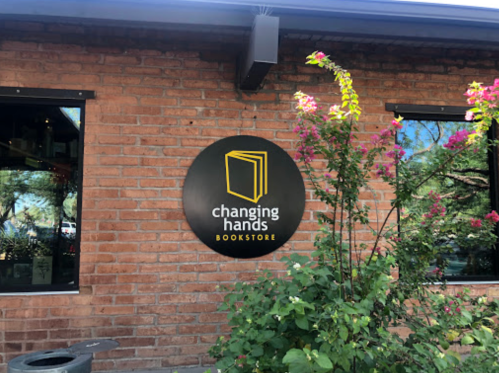 Sign for "Changing Hands Bookstore" on a brick wall, surrounded by green plants and flowers.